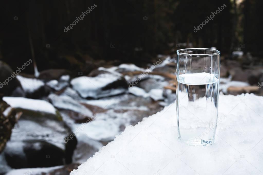 A transparent glass glass with drinking mountain water stands in the snow against a background of a clean mountain river and a forest in winter.