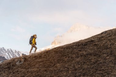 Bir backpacker büyük kürk şapka ve eldiven sırtında bir sırt çantası ile bulutlar epik kayalıklardan çerçevede yokuş yukarı gidiyor. Her mevsim dağ turizm kavramı