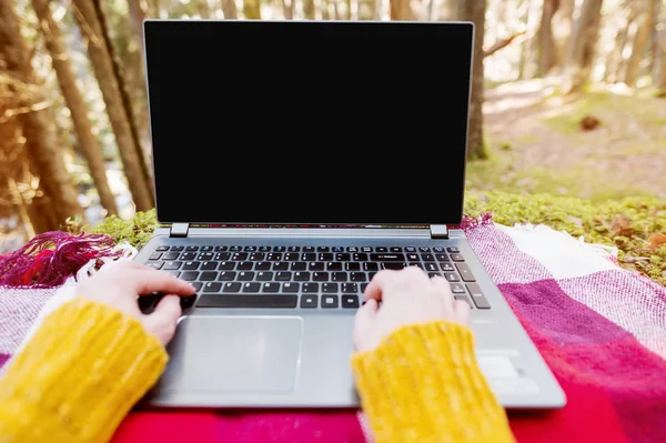 A mock-up image of a womans hand using and typing on a laptop with a blank black desktop on a plaid grass lying in the forest. The concept of freelancing in freedom from a permanent place of work and