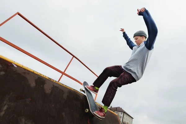 Un patinador adolescente con sombrero hace un truco de Rocks en una rampa en un parque de skate contra un cielo nublado y una zona para dormir. El concepto de estilo urbano en el deporte — Foto de Stock