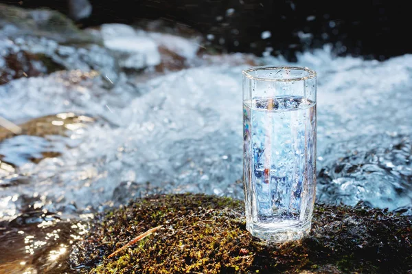 A transparent glass glass with drinking mountain water stands in the moss stone on sun beame against a background of a clean frost mountain river. The concept of drinking mountain drinking mineral