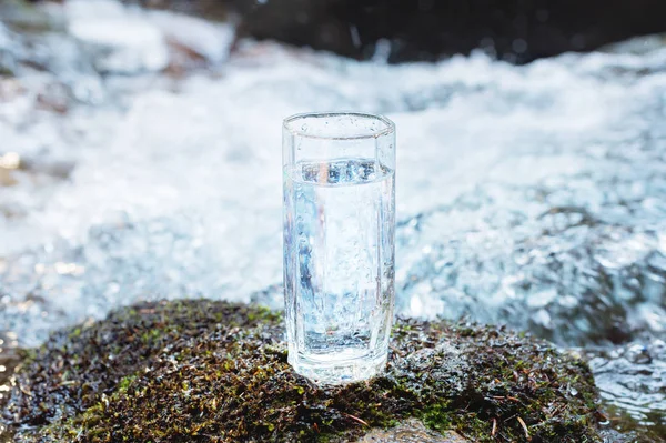 A transparent glass glass with drinking mountain water stands in the moss stone on sun beame against a background of a clean frost mountain river. The concept of drinking mountain drinking mineral