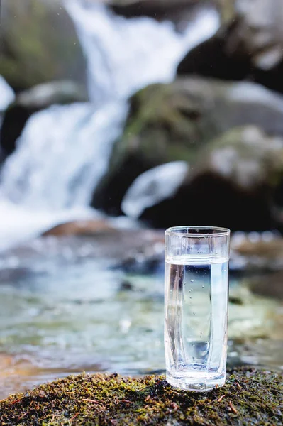 Un vetro trasparente con acqua potabile di montagna si trova nella pietra muschio sul raggio di sole sullo sfondo di un fiume di montagna gelo pulito. Il concetto di bere minerale di montagna — Foto Stock