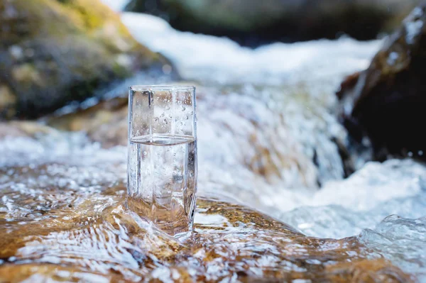 Un bicchiere di vetro con acqua potabile pura di montagna sorge su una roccia nel corso di un fiume di montagna sullo sfondo di cascate ribollenti e cascate di un fiume di montagna. Il concetto di puro — Foto Stock