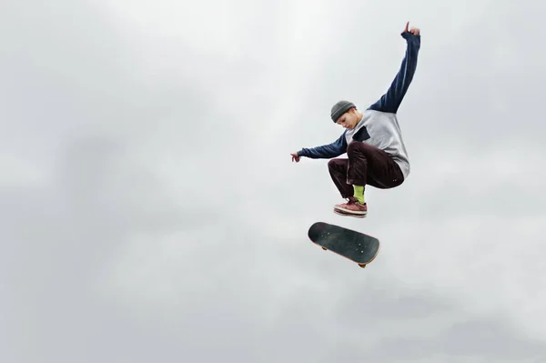 Nastolatek skater w kapeluszu i bluzę załatwia w powietrzu z flip zarządu przeciwko zachmurzone niebo szare. Skater jest odizolowana od innych obiektów przed niebo — Zdjęcie stockowe