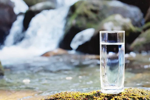 Un vetro trasparente con acqua potabile di montagna si trova nella pietra muschio sul raggio di sole sullo sfondo di un fiume di montagna gelo pulito. Il concetto di bere minerale di montagna — Foto Stock