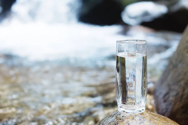 Un vetro trasparente con acqua minerale del fiume di montagna sorge su una pietra accanto al torrente del fiume di montagna — Foto Stock