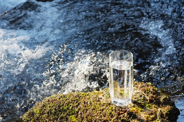 Un vetro trasparente con acqua potabile di montagna alla luce del sole si trova nella pietra muschio sul raggio di sole sullo sfondo di un fiume di montagna gelo pulito. Il concetto di bere in montagna — Foto Stock