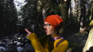 Bir etkin sağlıklı hippi kız fotoğrafçı elleri bir kamera ile ormanda yürür. Kız resimleri alıyor. Düşük anahtar 60 Fps ağır çekim. Üç eksenli antimikrobiyal GoPro 6 siyah