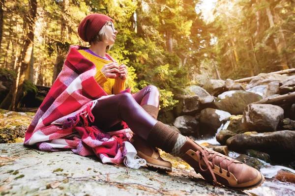 Uma menina bonita hipster com uma xadrez xadrez em seus ombros em um chapéu e um suéter amarelo e óculos com uma caneca de café em suas mãos olha para um rio de montanha sentado em uma rocha em uma floresta de pinheiros. O — Fotografia de Stock