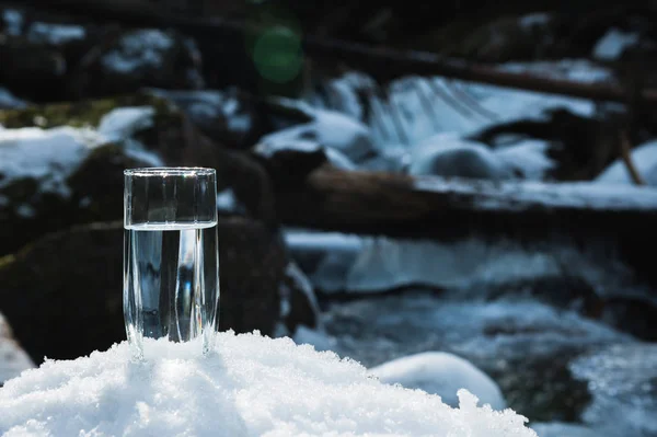 Un bicchiere di vetro trasparente con acqua potabile di montagna sorge nella neve sullo sfondo di un fiume di montagna gelo pulito in inverno . — Foto Stock