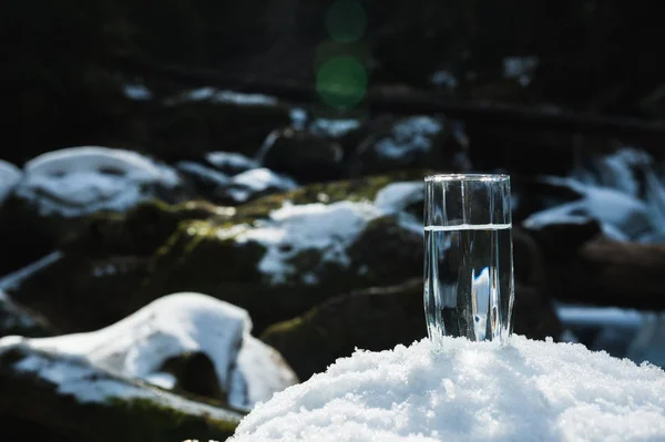 Un bicchiere di vetro trasparente con acqua potabile di montagna sorge nella neve sullo sfondo di un fiume di montagna gelo pulito in inverno . — Foto Stock