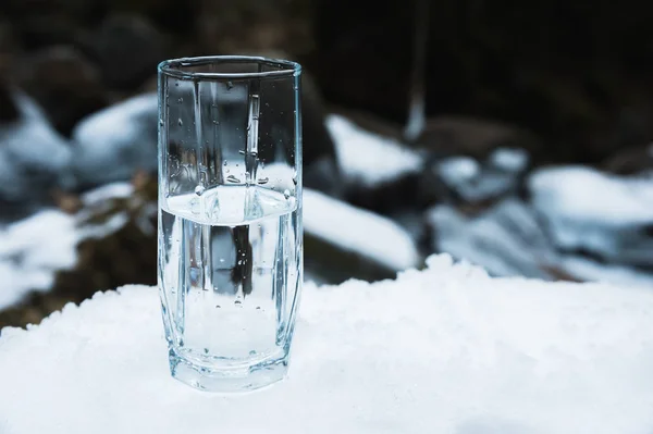A transparent glass glass with drinking mountain water stands in the snow against a background of a clean frost mountain river in winter.