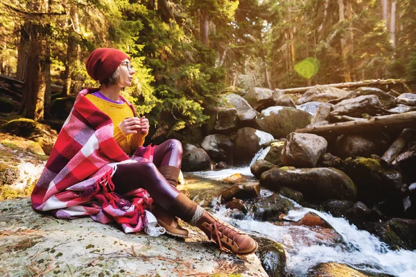 모자와 노란색 스웨터에 그녀의 어깨에 격자 무늬 격자 무늬와 예쁜 hipster 소녀와 그녀의 손에 커피의 낯 짝으로 안경 소나무 숲에 바위에 앉아 산 강에 보인다. 는 — 스톡 사진
