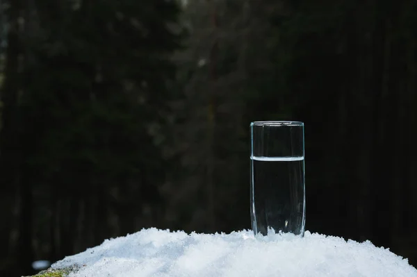 Een transparant glas glas met drinkwater van de berg staat in de sneeuw tegen een achtergrond van een bos in de winter. — Stockfoto