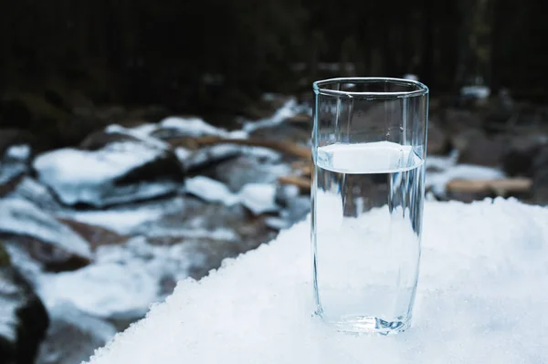 Un bicchiere di vetro trasparente con acqua potabile di montagna sorge nella neve sullo sfondo di un fiume di montagna gelo pulito in inverno . — Foto Stock