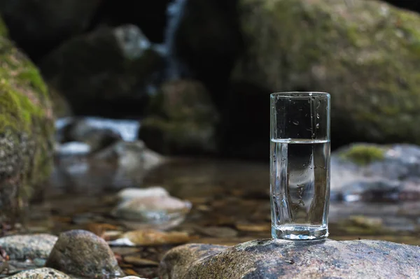 Un vetro trasparente con acqua minerale del fiume di montagna sorge su una pietra accanto al torrente del fiume di montagna — Foto Stock