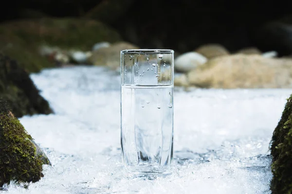Transparent glass glass with drinking mountain water in winter stands on an icy crust against the background of a clean, frosty river bank. The concept of drinking mountain drinking mineral water and