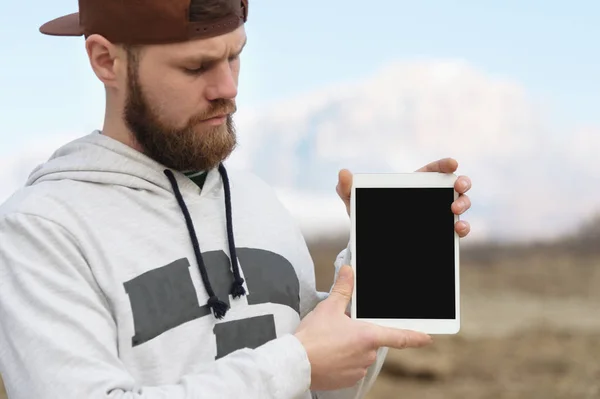 Close-up Retrato de um hipster em uma tampa marrom ao ar livre segura um pc tablet branco em suas mãos. Um homem barbudo olha para o tablet. borrão de fundo — Fotografia de Stock