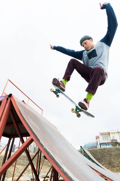 Un adolescente patinador con sombrero hace un truco con un salto en la rampa. Un patinador vuela en el aire —  Fotos de Stock