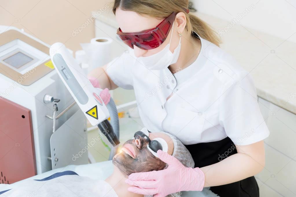 The cosmetician girl in goggles makes the procedure of carbon peeling with the help of a cosmetology laser. Carbon face peeling procedure. Laser pulses clean skin of the face. Hardware cosmetology