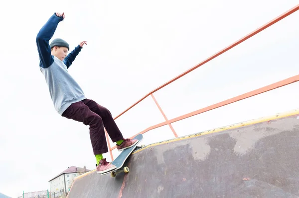 Adolescente patinador em uma camisola com capuz e jeans desliza sobre um corrimão em um skate em um parque de skate — Fotografia de Stock