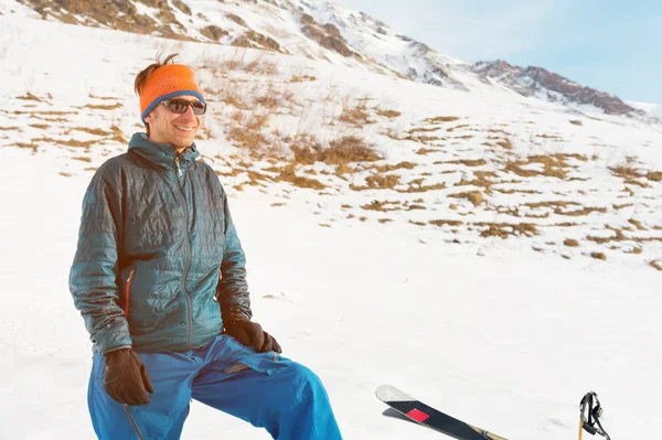 Um turista de jaqueta e óculos de sol com luvas fica nas montanhas do Cáucaso ao lado dos esquis e ri — Fotografia de Stock