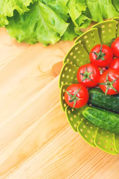 Färska grönsaker gurkor och tomater i en grön skål på ett träbord bredvid gröna salladsblad. Ovanifrån i en tabell. Vegetarisk kost. Hälsosam kost och livsstil — Stockfoto