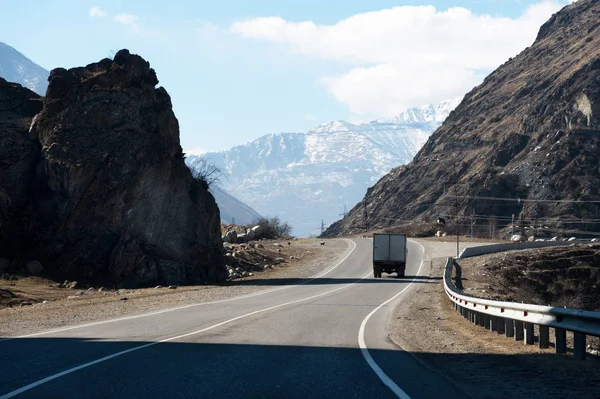 Coche de carga en una carretera de montaña junto a las rocas con un cielo azul y montañas en el fondo. El concepto de empresas de transporte y el transporte de mercancías y mercancías a lugares de difícil acceso — Foto de Stock