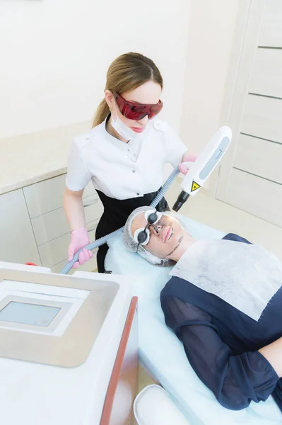 A menina cosmetician em óculos faz o procedimento do descascamento de carbono com a ajuda de um laser cosmetology. Procedimento de descascamento da cara de carbono. Pulsos laser pele limpa do rosto. Cosmetologia de hardware — Fotografia de Stock