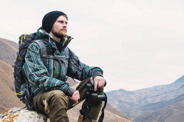 Yorgun sakallı hippi ellerinde dürbün ile bir taş dağları arasında oturur ve mesafe içine bakıyor