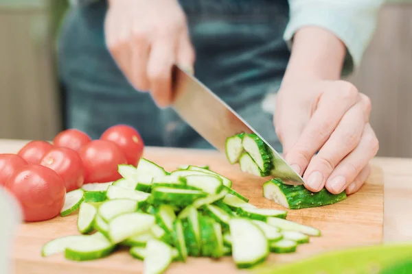 Primer plano de las manos femeninas cortadas en pepinos cortados frescos en una tabla de cortar de madera junto a los tomates rosados. El concepto de cocina vegetariana casera y alimentación saludable y estilo de vida — Foto de Stock