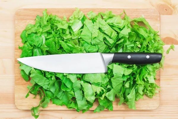 El primer plano sobre una mesa de madera es una tabla de cortar de madera en la que se encuentran las hojas de lechuga picadas y en ellas se encuentra un cuchillo de corte grande. El concepto de comida vegetariana autococinada en casa. Saludable — Foto de Stock