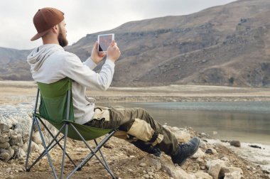 Bir kap içinde sakallı bir hipster katlanır sandalye yüksek dağ Gölü kıyısında oturur ve bir tablet üzerinde bir fotoğrafı çeker. Sakallı bir blogger doğadan yayın yapmıyor. Seyahat blogcular kavramı