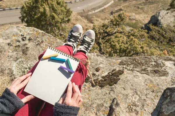 View from the first person on the female lap is a notebook with chalks for drawing pastels. Female hands holding a notebook in nature. A girl sitting on a cliff near a cliff is going to paint a pastel