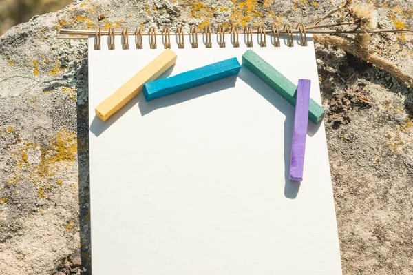 Close-up artwork for the designer. A blank notepad for drawing pastel on which lay an artistic pastel crayons color chalks of purple green blue and yellow lies on a rock in nature outdoors. The — Stock Photo, Image