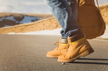 Yaz günbatımı asfalt üzerinde yürürken, sarı Ayakkabı ve mavi jeans mens bacaklarda asfalt gidin. Vintage sırt çantası yanında. Otostop kavramı