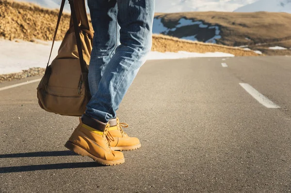 Caminando en el atardecer de verano sobre el asfalto, las piernas de hombre en zapatos amarillos y pantalones vaqueros azules van a lo largo del asfalto. Junto a la mochila vintage. Concepto de autoestop — Foto de Stock