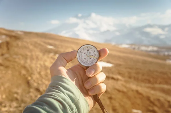 Een mans hand bezit een hand-held kompas tegen de achtergrond van bergen en heuvels bij zonsondergang. Het concept van reizen en navigatie in open gebieden — Stockfoto
