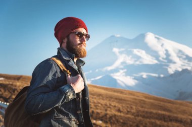Düşünceli bir şık hipster güneş gözlüğü sakallı ve gün batımında Kafkasya'da karla kaplı Elbruz Dağı arka plan üzerinde bir sırt çantası ile bir şapka portre