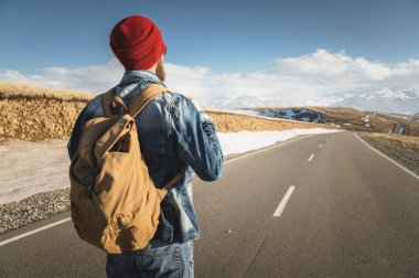 Kırmızı bir şapka ve kot ceket ve kot pantolon ile güneş gözlüğü takıyor bir eski moda vintage sırt çantası ile sakallı bir hipster bir kırsal asfalt dağ yolda duruyor ve karla kaplı görünüyor