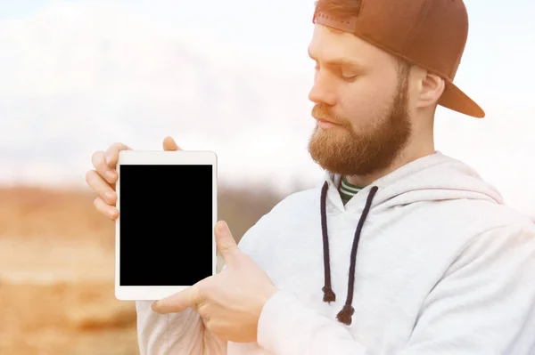 Close-up Retrato de um hipster em uma tampa marrom ao ar livre segura um pc tablet branco em suas mãos. Um homem barbudo olha para o tablet. borrão de fundo — Fotografia de Stock