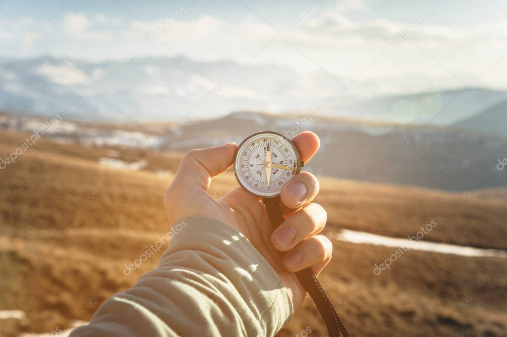 a mans hand holds a hand-held compass against the backdrop of mountains and hills at sunset. The concept of travel and navigation in open areas