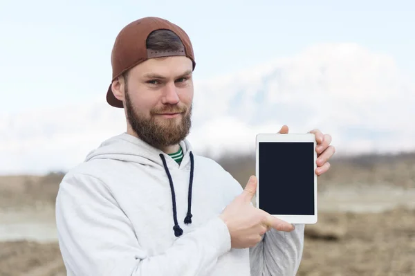 Retrato de close-up de um hipster sorridente em uma tampa marrom ao ar livre segurando um computador tablet branco com um recorte em branco para o designer. Um homem barbudo olha para o tablet. borrão de fundo . — Fotografia de Stock
