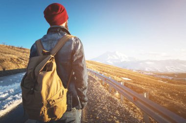 Kırmızı bir şapka ve kot ceket ve kot pantolon ile güneş gözlüğü takıyor bir eski moda vintage sırt çantası ile sakallı bir hipster bir kırsal asfalt dağ yolda duruyor ve karla kaplı görünüyor