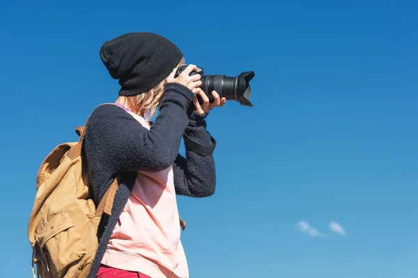 Портрет стильної дівчини хіппі в капелюсі і з рюкзаком, який фотографує її на камеру DSLR на відкритому повітрі проти блакитного неба в сонячний день. Концепція фотографії в подорожах — стокове фото