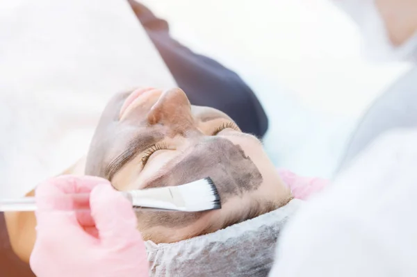 Το cosmetologist σε ροζ γάντια με ένα πινέλο ισχύει μια μάσκα άνθρακα για peeling στο πρόσωπο του ένα νεαρό κορίτσι σε ένα δωμάτιο κοσμετολογία. Η έννοια της κοσμετολογίας υπηρεσίες και self-care. Η έννοια της — Φωτογραφία Αρχείου