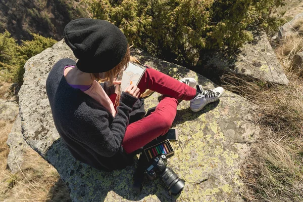 Портрет дівчини-фотографа-гіпстера в капелюсі і сонцезахисних окулярах малює пастель в записнику, що сидить на скелі, відкритому в горах. Концепція вільного фрілансера — стокове фото