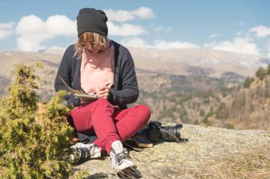 Güneş gözlüğü ve kayada bir açık mavi gökyüzü karşı dağlarda oturan bir şapka giyen bir hipster kız portresi. Freelancer seyahat tasarımcısında bir kalem bir defterde çizer.