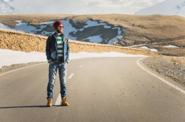 Güneş gözlüğü vintage bir sırt çantası ile şık bir sakallı hippi güneşli bir günde bir ülke yol asfalt üzerinde duruyor. Otostop ve hiking kavramı